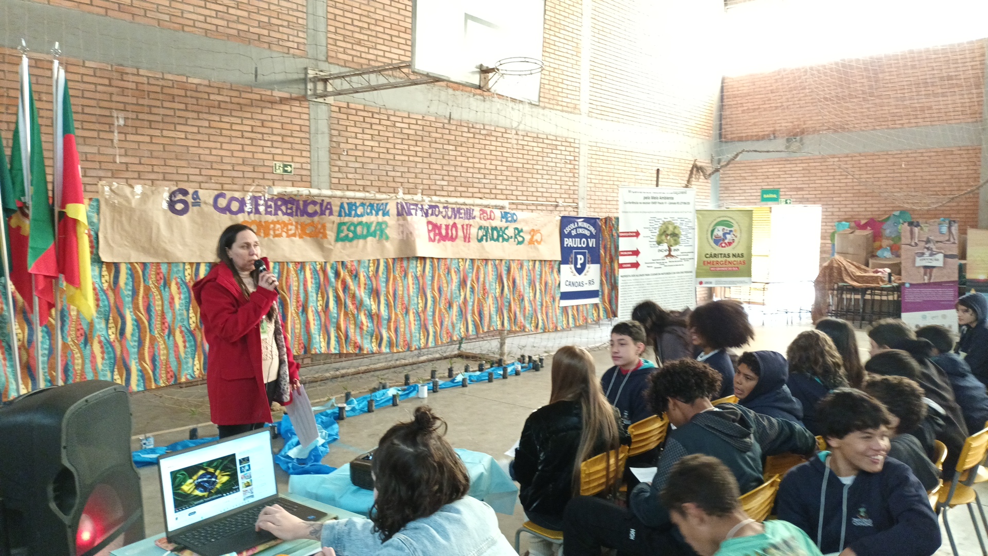 A diretora Maristela de Oliveira Borsoi (de vermelho) fala na abertura da Conferência InfantoJuvenil pelo Meio Ambiente na EMEF Paulo VI. À direita, ao fundo, o cartaz verde do Projeto Cárias nas Emergências